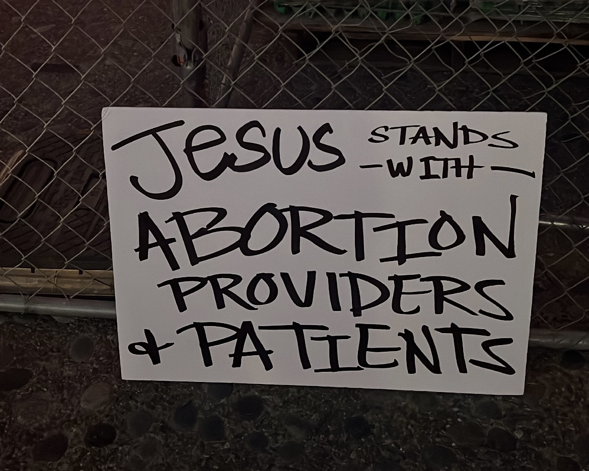 A white protest sign with "Jesus stands with abortion providers and patients" written in thick, black permanent marker. The sign is leaning against a chain-link fence at dusk.